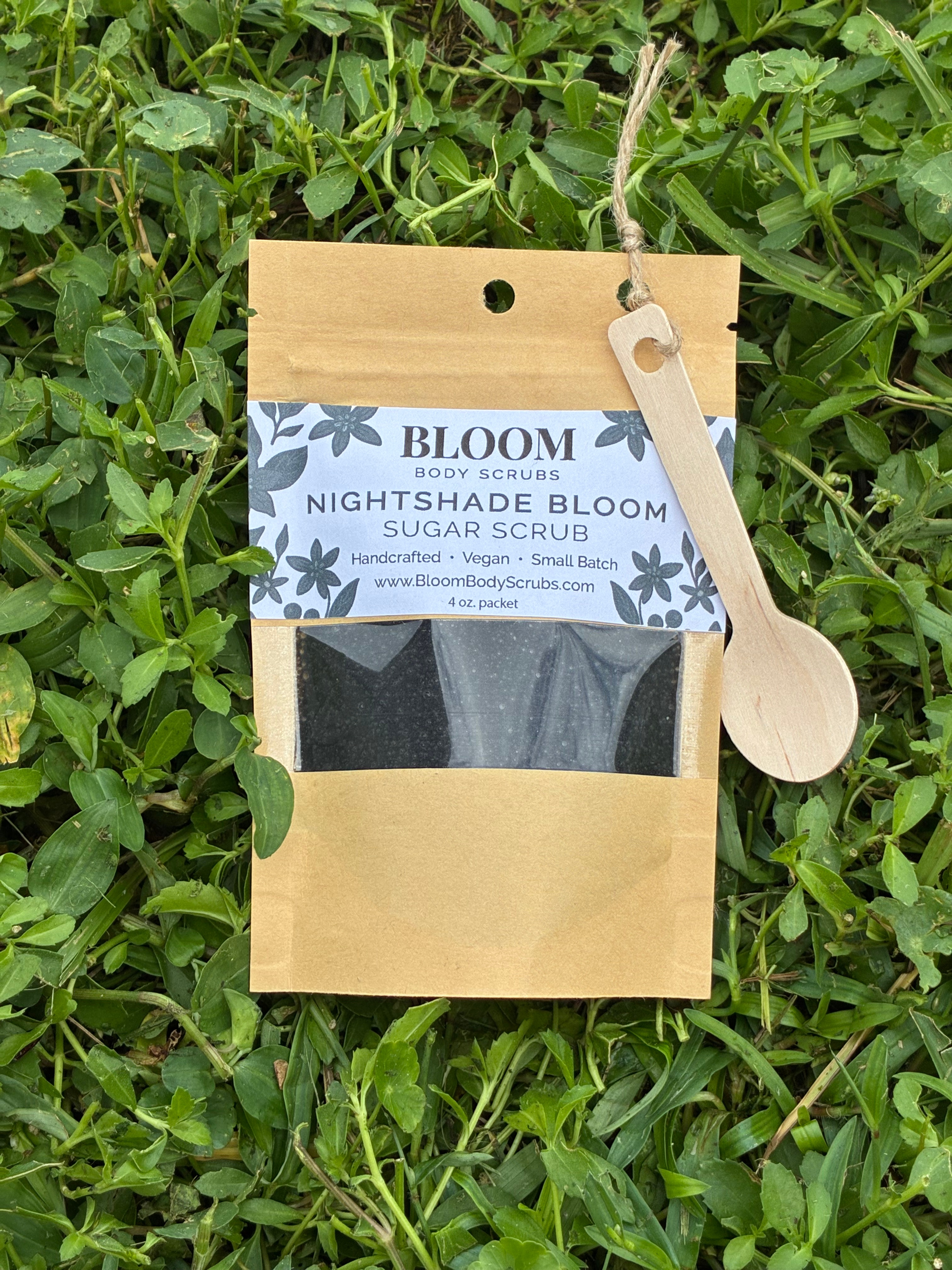 Bloom's Nightshade Bloom Body Scrub shown in Kraft paper resealable packet with a small wooden spoon attached with twine with vibrant green grass in the background.