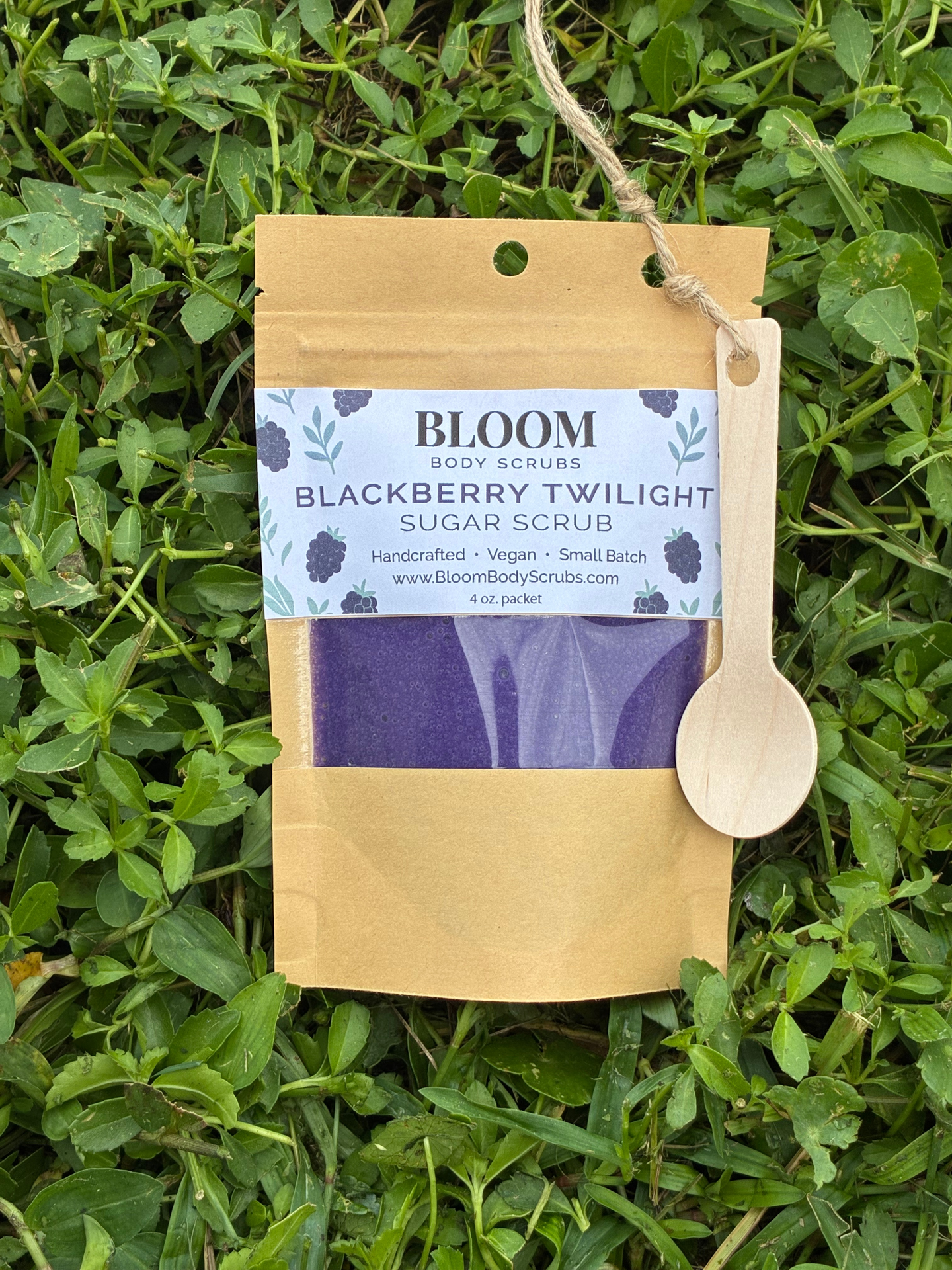 Bloom's Blackberry Twilight Body Scrub shown in Kraft paper resealable packet with a small wooden spoon attached with twine with vibrant green grass in the background.