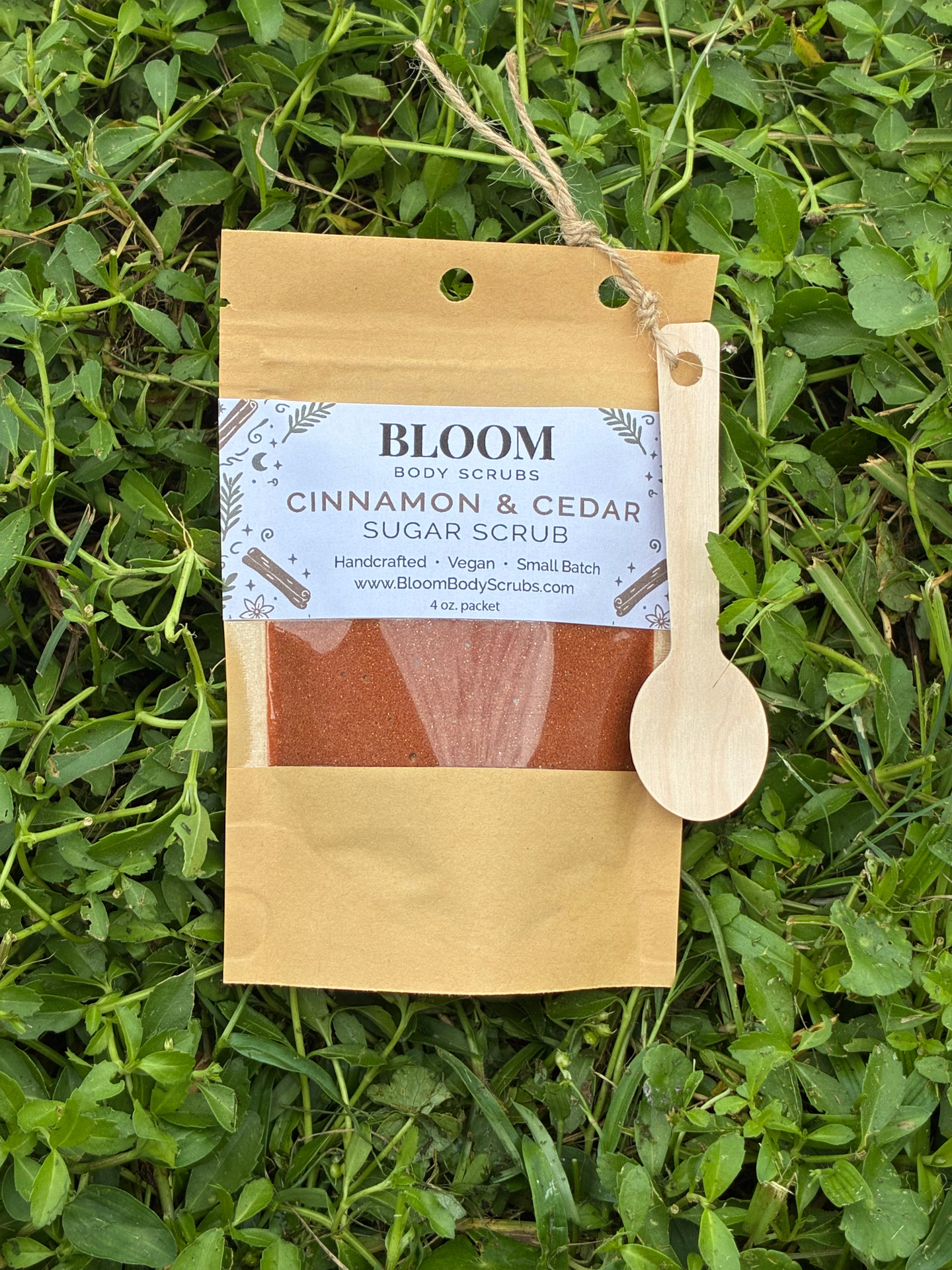 Bloom's Cinnamon & Cedar Body Scrub shown in Kraft paper resealable packet with a small wooden spoon attached with twine with vibrant green grass in the background.